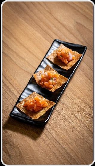 Três canapés com salmão picado por cima, alinhados na travessa preta