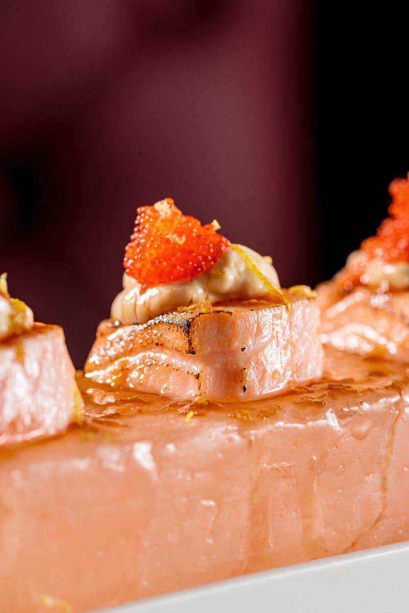 Quatro cubos de salmão com creme e ovas alaranjadas por cima, alinhados sobre a pedra de sal rosa