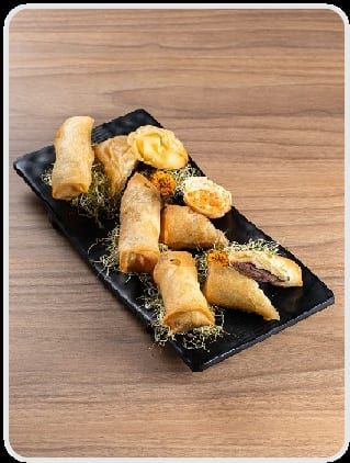 Rolinhos dourados e crocantes deitados na travessa preta, com crispy verde ao redor