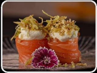 Dois joys enrolados em salmão, com creme e fios dourados de alho-poró frito no topo, e flores ao lado
