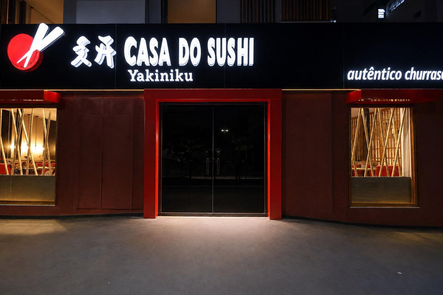Fachada frontal noturna com o letreiro Casa do Sushi Yakiniku aceso sobre a porta vermelha e as vitrines de bambu