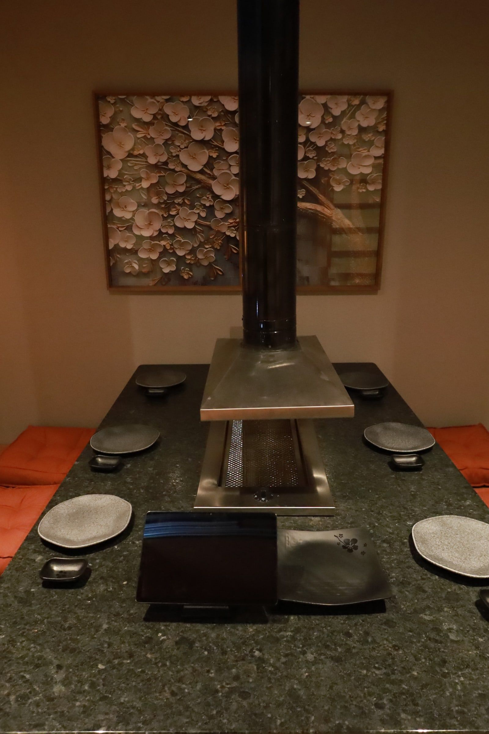 Sala privativa em tatame com quadro de cerejeira em relevo, coifa preta e mesa rebaixada de grelha entre almofadas laranja