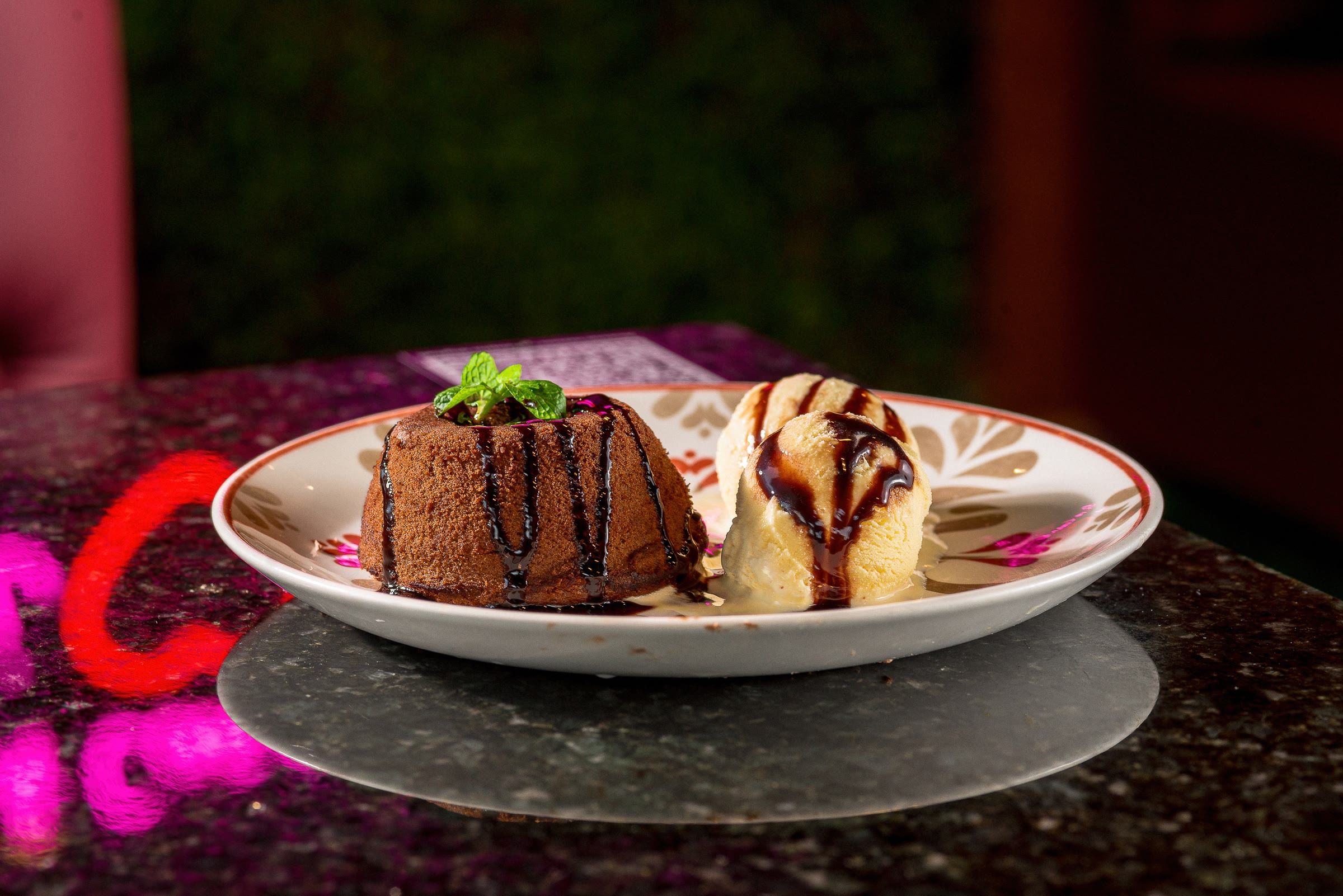 Bolo de chocolate com calda e sorvete em prato branco floral, sob o neon rosa do salão