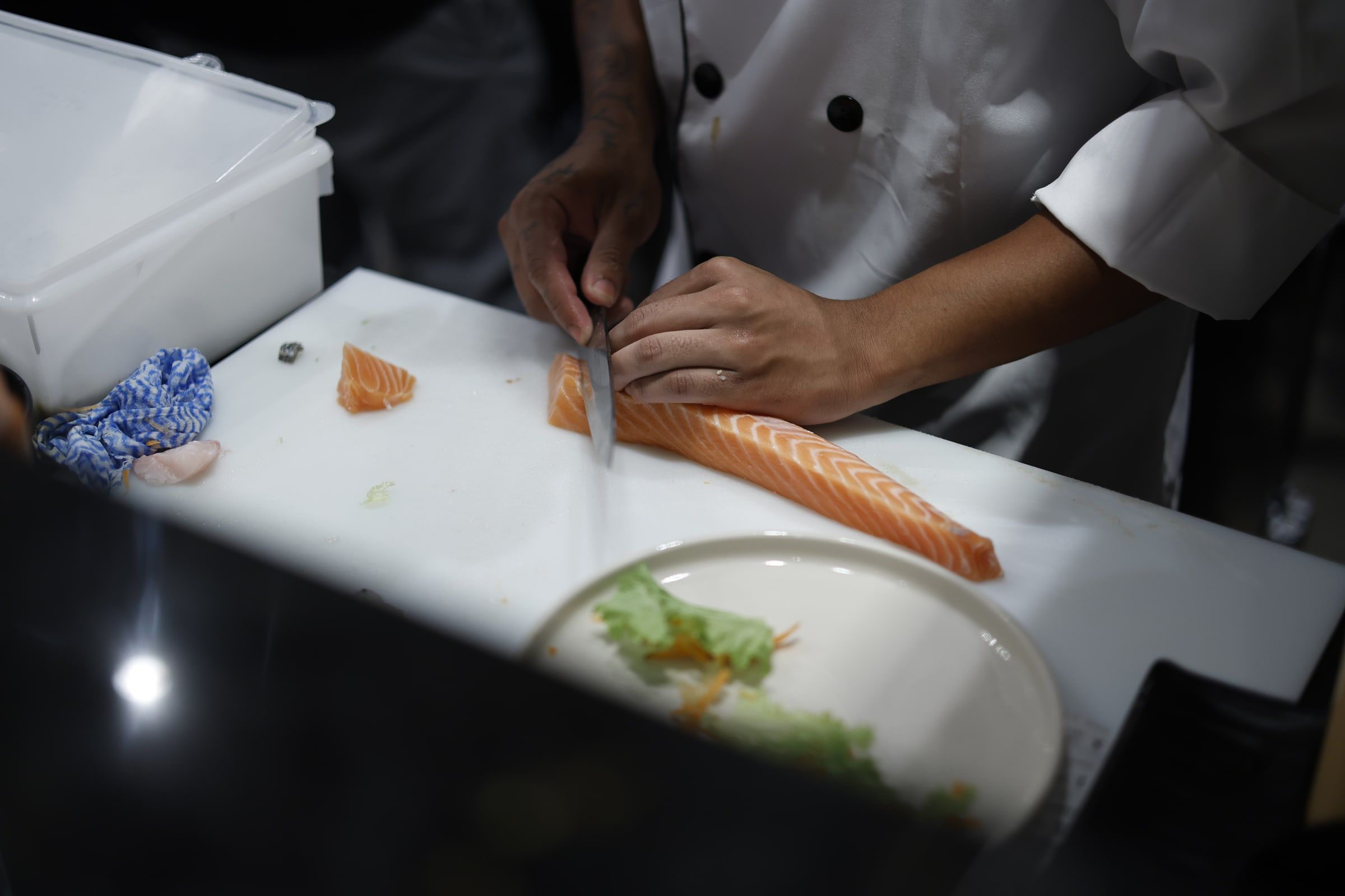 Mãos do sushiman fatiando um lombo de salmão com faca longa sobre a tábua branca do balcão