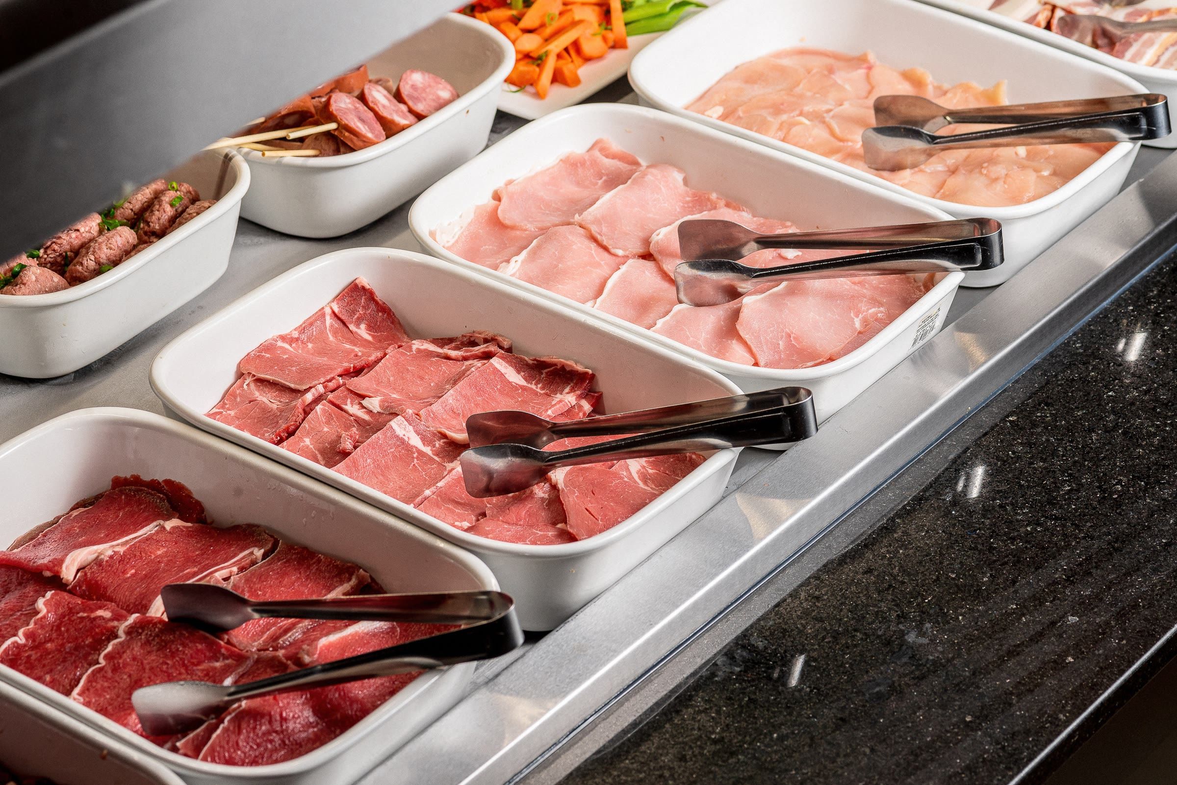 Bandejas brancas de carne crua fatiada e cubas de legumes coloridos, com pegadores pretos