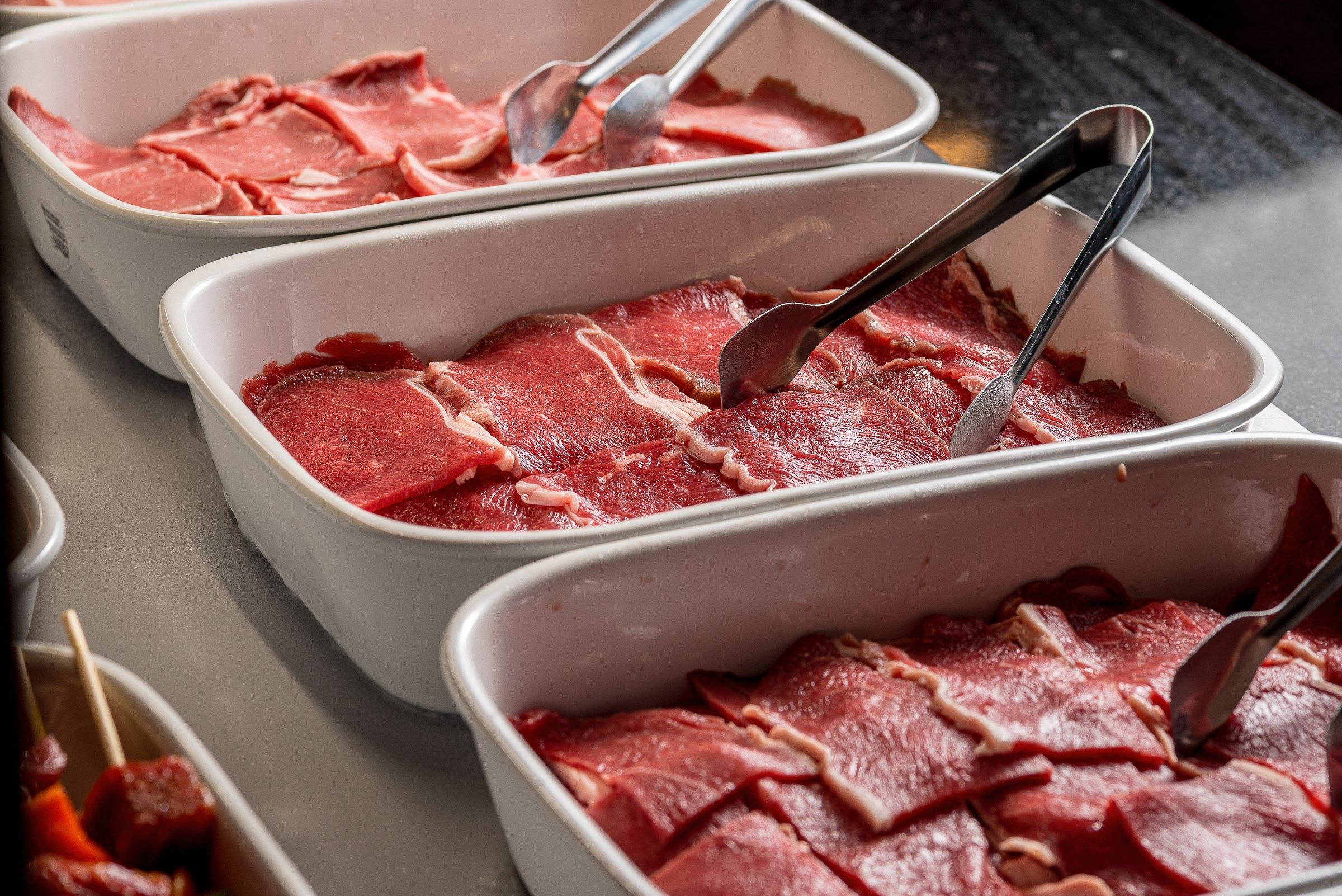 Cubas brancas com fatias finas de carne bovina crua e pegadores de inox no balcão de serviço