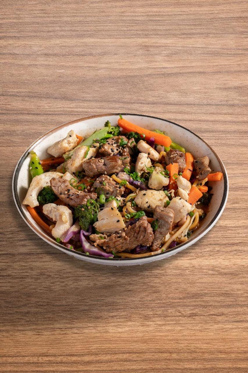 Yakissoba com tiras de carne, frango, brócolis e cenoura em tigela clara sobre mesa de madeira